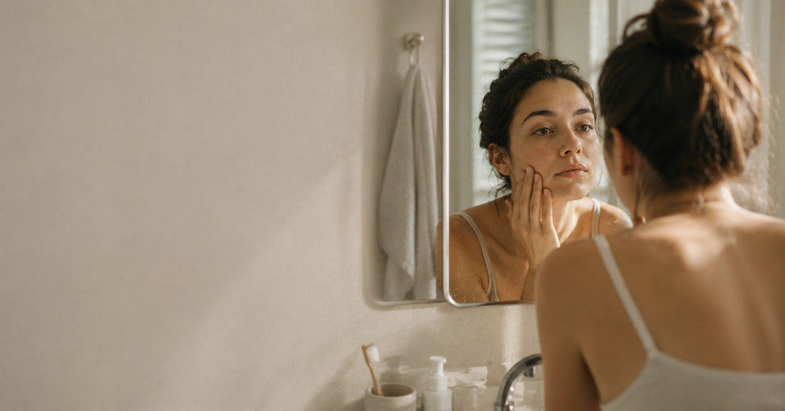 A woman looking closely at her reflection in a bathroom mirror after her morning skincare routine, representing the quiet frustration of skin that won't improve
